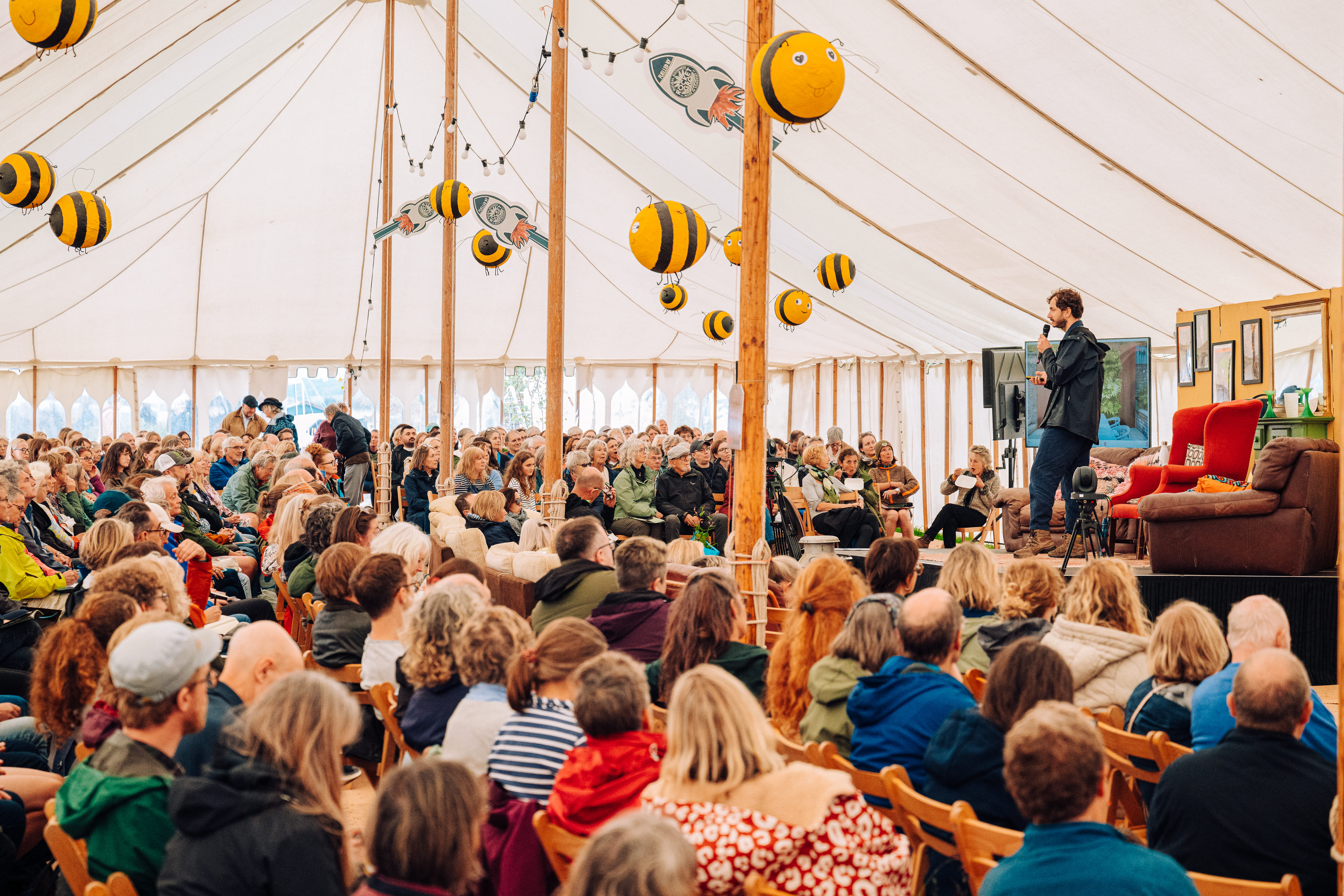 A festival in a field by a large lake. Children play with hula loops in the foreground as others sit on a picnic blanket in a circle. Behind them, several large tents have moderately dense crowds beside them. One of these tents is a sound stage.