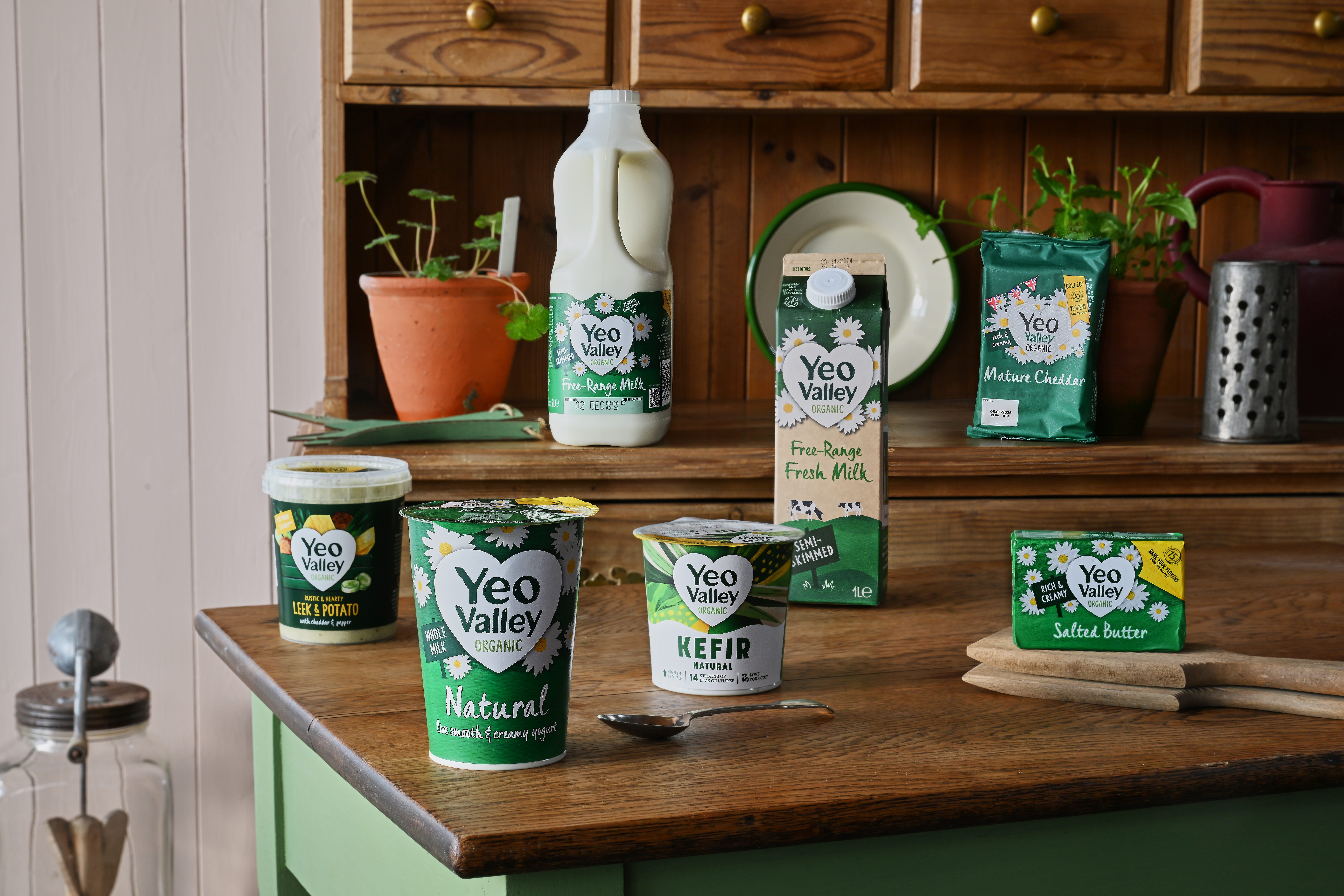A variety of Yeo Valley products are displayed on a wooden countertop. From left to right: Vanilla Ice Cream, Strawberry creamy bio live yoghurt, salted butter, natural creamy bio live yoghurt, Greek style natural yoghurt, Free-range fresh milk, Fruity favourites yoghurt, Kefir, Double cream, Mature cheddar and a little Yeo organic yoghurt pouch